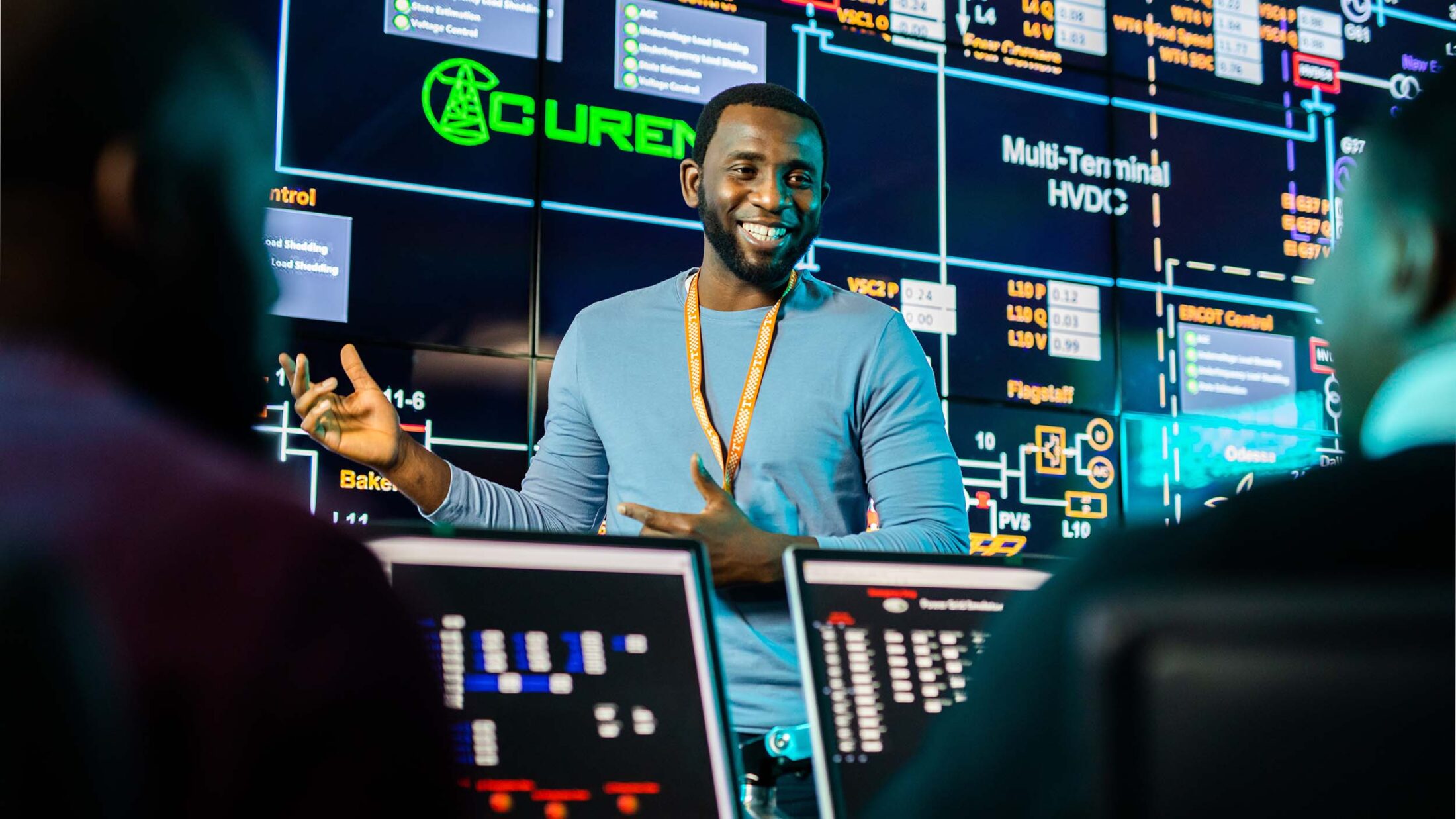 A man stands in front of a large screen and talks to two people using laptops.