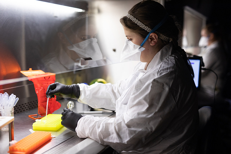 A woman wearing a white mask and lab coat uses a pipette while testing saliva for COVID-19.