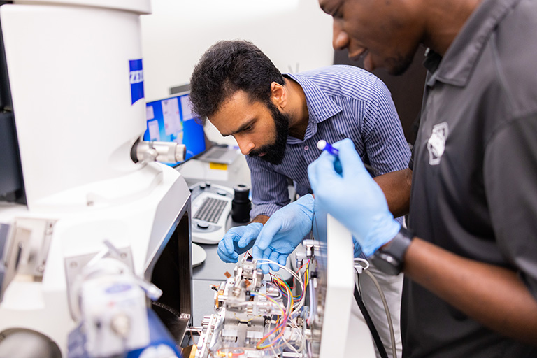 Two men wearing blue gloves work on the scanning electron microscope.