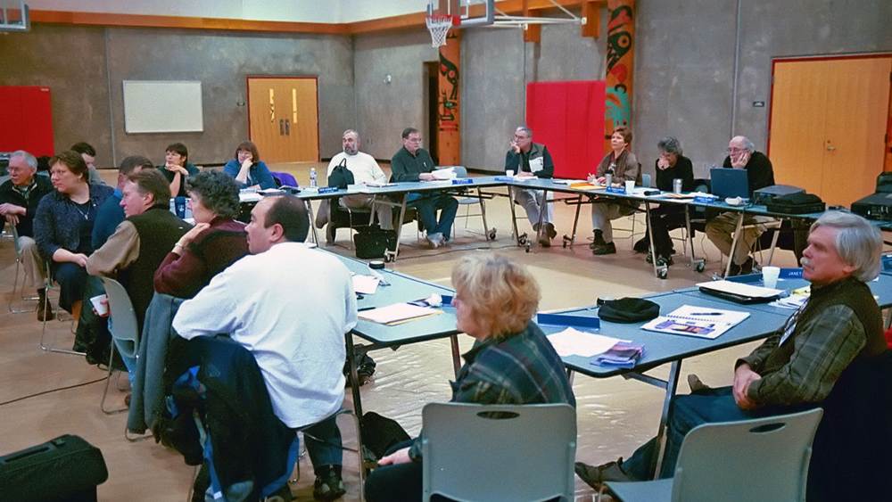 people sit in an auditorium at several tables arranged into a large square