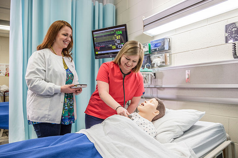 student and instructor standing over bed with nursing dummy