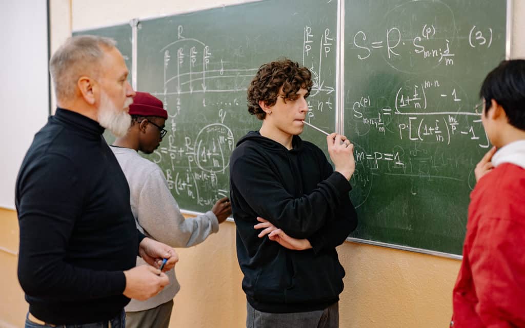 group of three students working on mathematic problems at chalkboard with teacher