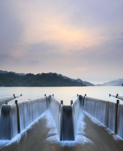 A symmetrical dam with flowing waterfalls under a cloudy sky, surrounded by lush green hills and a tranquil lake.