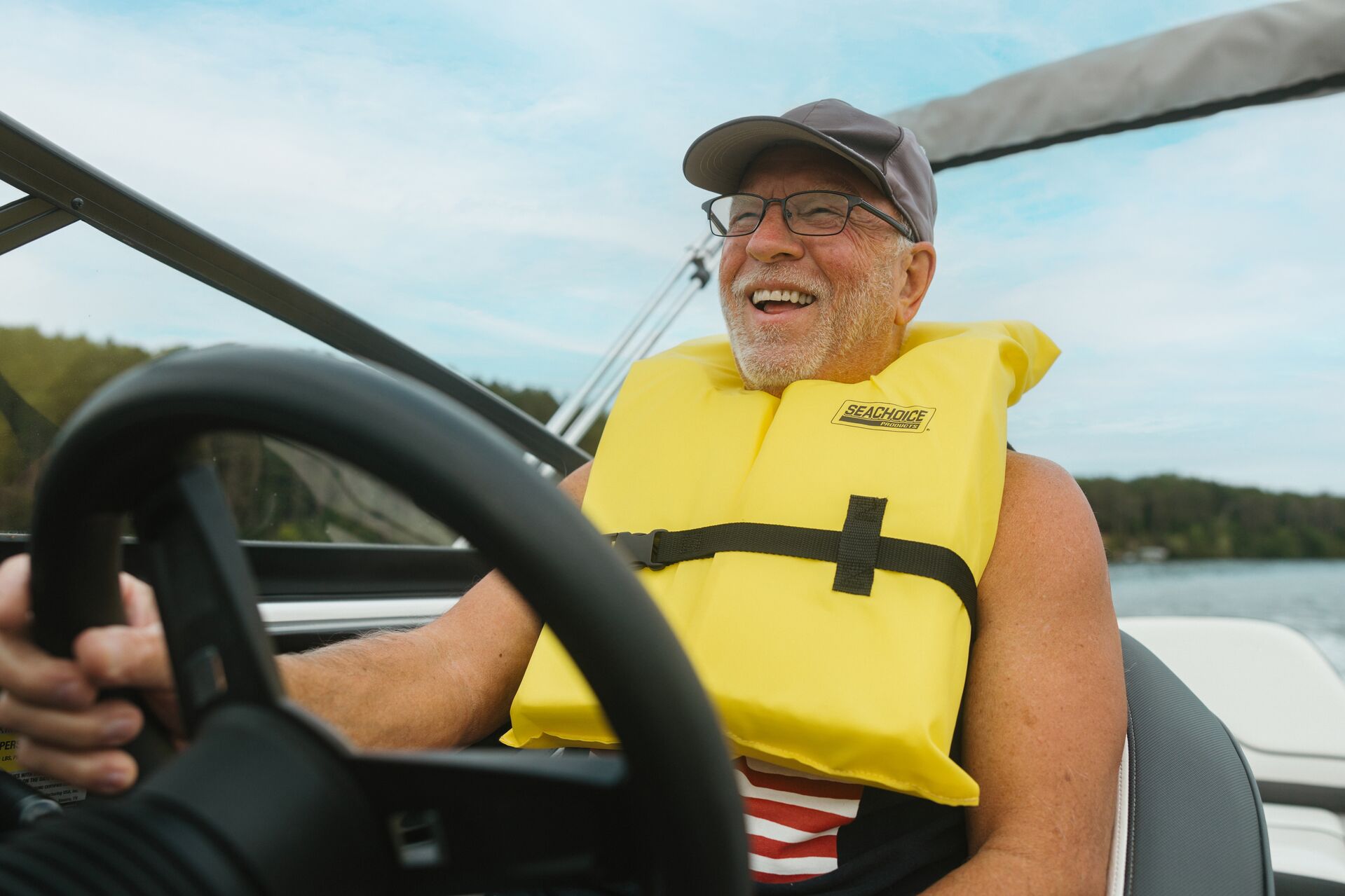 A man smiles while driving a boat and wearing a yellow life vest. 