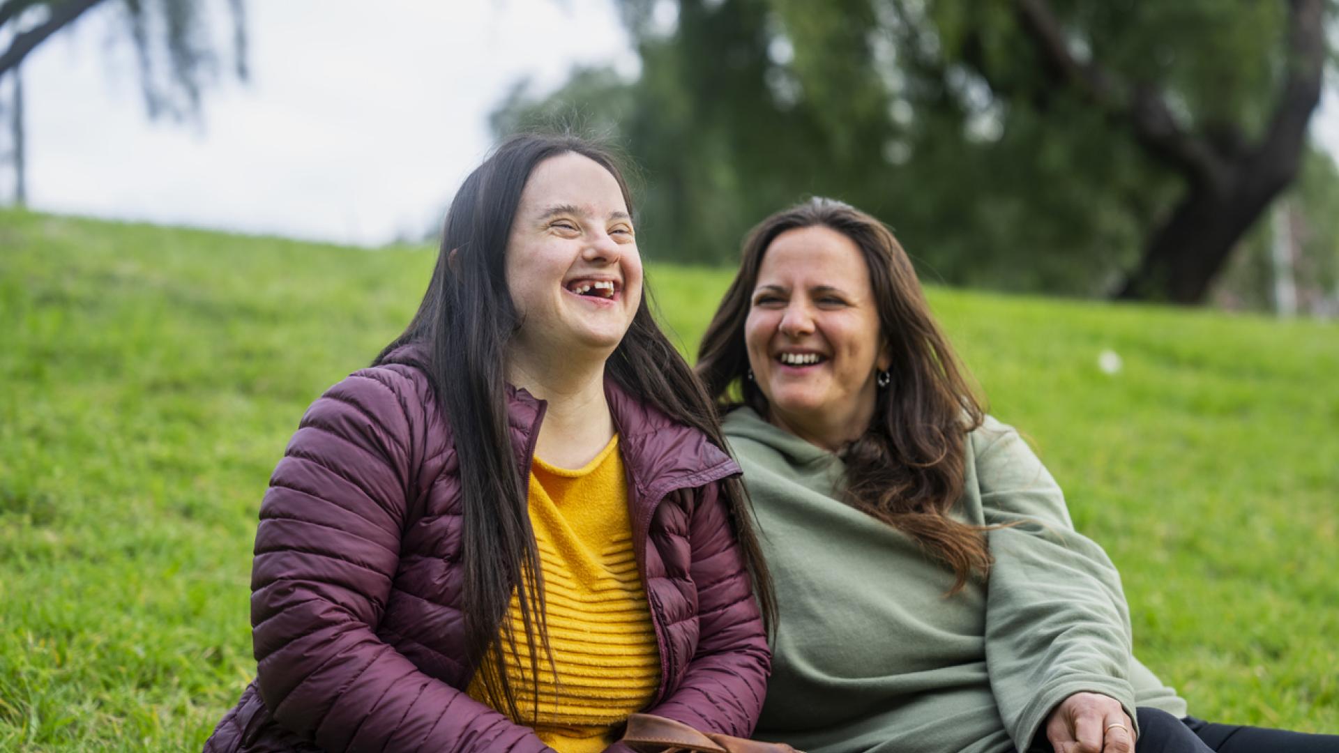 Two women sitting together on a grassy hill, enjoying a moment of laughter. The woman on the left, with Down syndrome, wears a purple jacket and yellow sweater, while the woman on the right, wearing a green hoodie, smiles warmly. Trees and a soft, overcast sky are visible in the background.