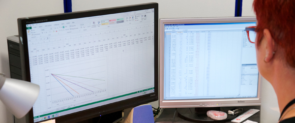 A person with red hair sat at a desk facing two computer screens. Once screen has a graph on it and the other has a spreadsheet