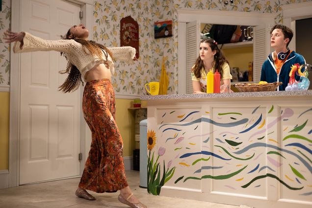 Photo: Summer Sage, played by Michaela Lazarou (CFA’24) does a unique interpretative dance as she holds her arms out and throws her head back towards the ceiling. To her right, behind a colorfully painted kitchen island, stand Candy Baker, played by Katie McRae (CFA’23), and Lil Bub, played by Aidan Close (CFA’23). Both look on in shock and awe.