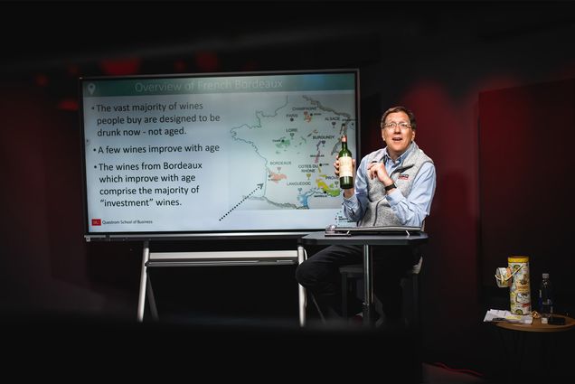 Photo: Professor Jay Zagorsky, a white man wearing glasses holding a bottle of wine, lectures on a stage in front of a whiteboard depicting Bordeaux France.