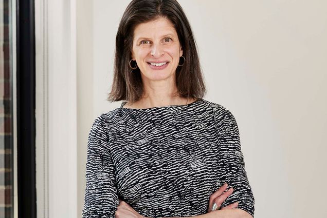 Photo: Dr. Emily Rothman, a white woman with shoulder-length brown hair and wearing a black and white half-sleeved dress, stands and poses with arms crossed. She smiles as she stands next to a large window in a brightly lit space.