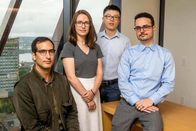 Photo: Four individuals take a group shot. From the left, a man with a dark green bomber jacket and glasses. Next to him, standing, a woman with shoulder length brown hair and glasses has her arms clasped. Behind her, a man with a blue button up, glasses, and short dark hair stands. And on the right, a man with short dark hair, glasses, a button up and gray slacks sits on a table. A huge window stands behind them.