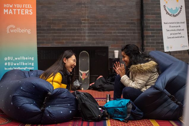 Photo: Two woman laugh joyfully at the Wellbeing Experience, an event curated to help destress during college finals. They sit on human sized beanbags wearing their winter coats. Behind them are informational signs about the event.