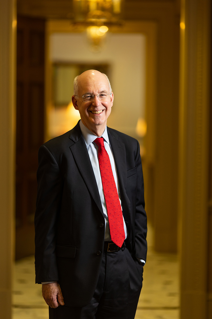 Photo: Kenneth Freeman, a white man wearing glasses, a light blue collared shirt, red tie, and black suit smiles and poses at 1 Silber Way in an elaborate hallway.