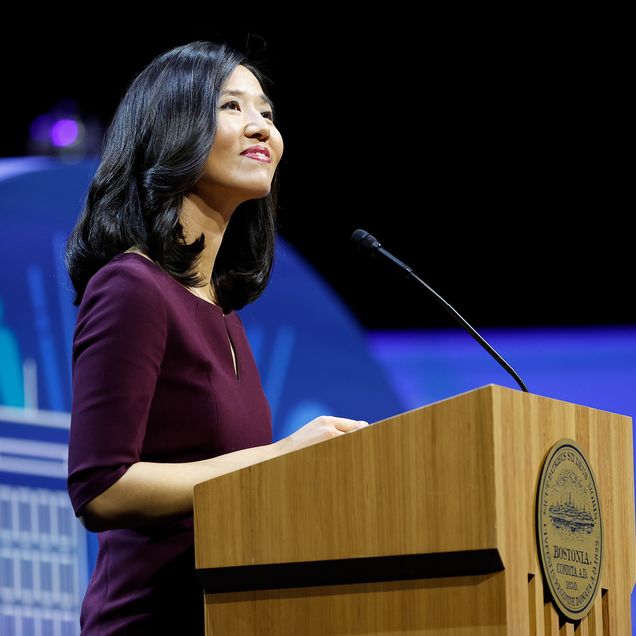 Photo: A picture of Boston Mayor Michelle Wu standing behind a podium. She is wearing a quarter-sleeve burgundy dress and smiling while looking into the crowd