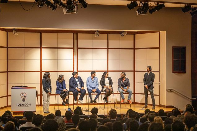 Photo: Five people sitting on stools on stage next to a podium that says "Idea Con." Two people stand on either side of them and a crowd is sitting in front of them