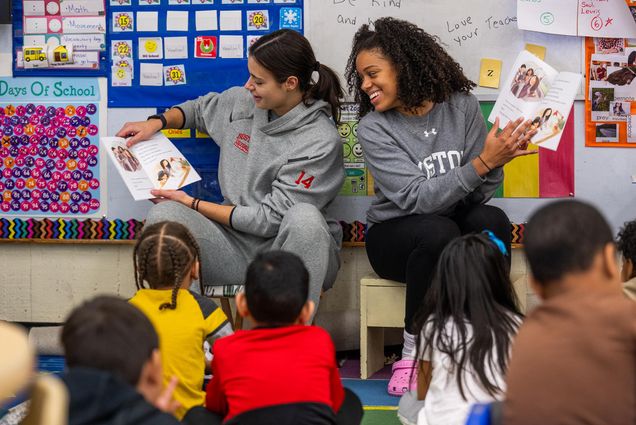 Photo: Two student athletes read to a group of kids in a classroom.