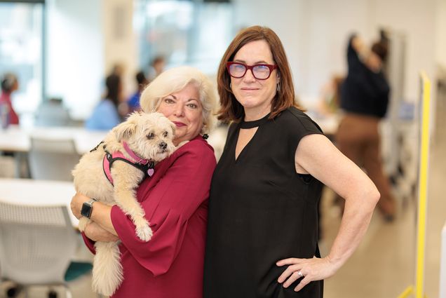 Photo: Two women standing side by side, one wearing red and holding a small white dog, the other wearing black with black glasses