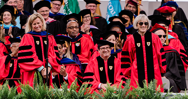 Photo: People standing on a stage lined with greenery in Boston University red academic robes with black trim and caps