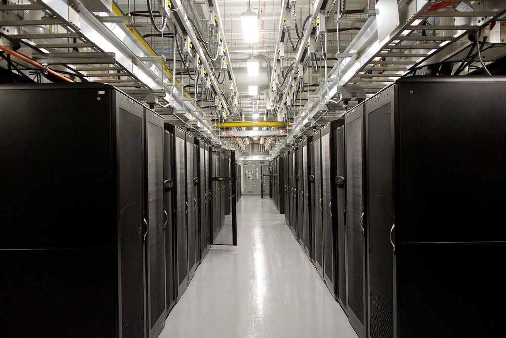 Photo: Interior of an AI data center, with large black server racks filling a gray room with steel cables and other wiring running along the ceiling