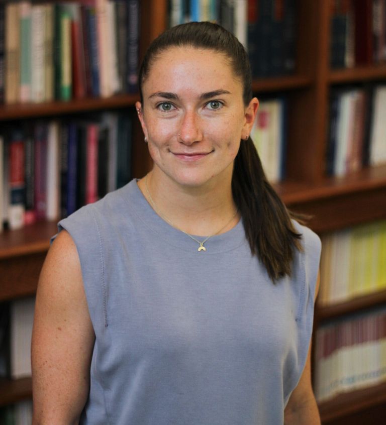 Malu Foley stands in front of a large bookcase and smiles at the camera