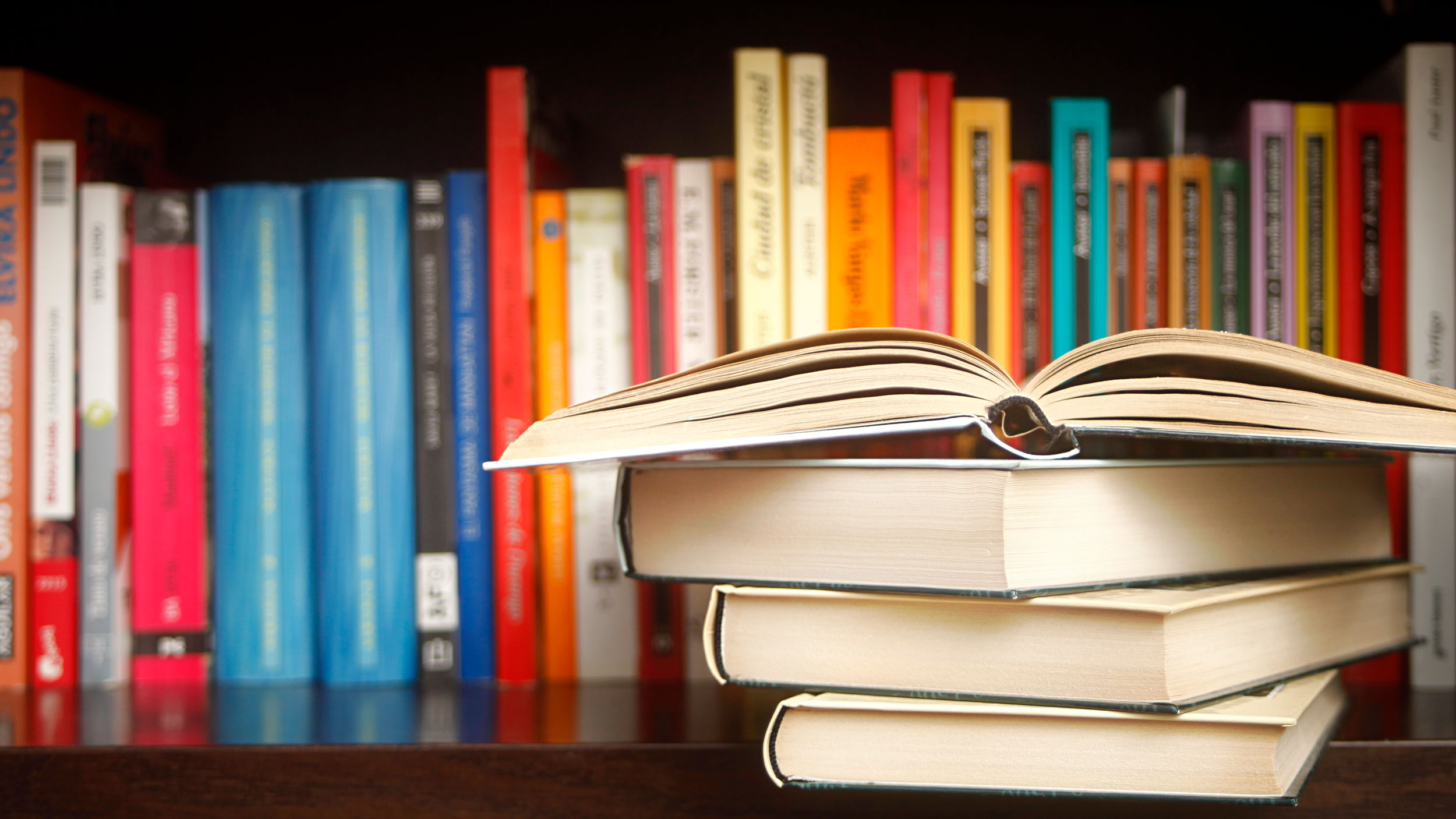 Row of books on a shelf, multicolored book spines, stack in the foreground.