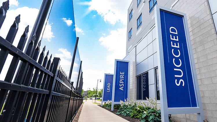 Path next to New Hall building on main campus with signs reading "Lead", "Aspire", "Succeed".