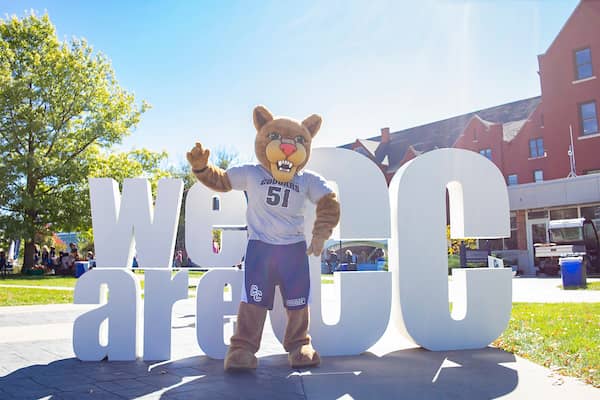 Scooter the Cougar standing outside on main campus in front of the We Are CC sign.