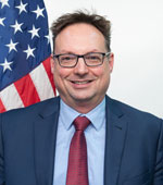 Headshot of Arron Helm. Arron has short hair and is wearing a blue suit and tie with glasses.