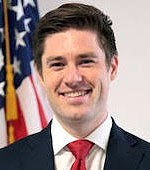 Headshot of Brett Prather. Brett has short brown hair and is wearing a black suit with a red tie.