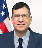 Headshot of Tom Howder. Tom has short dark hair and is wearing a blue suit and tie.