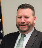 Headshot of Tom Lyman. He is wearing a dark suit with a sage green tie.