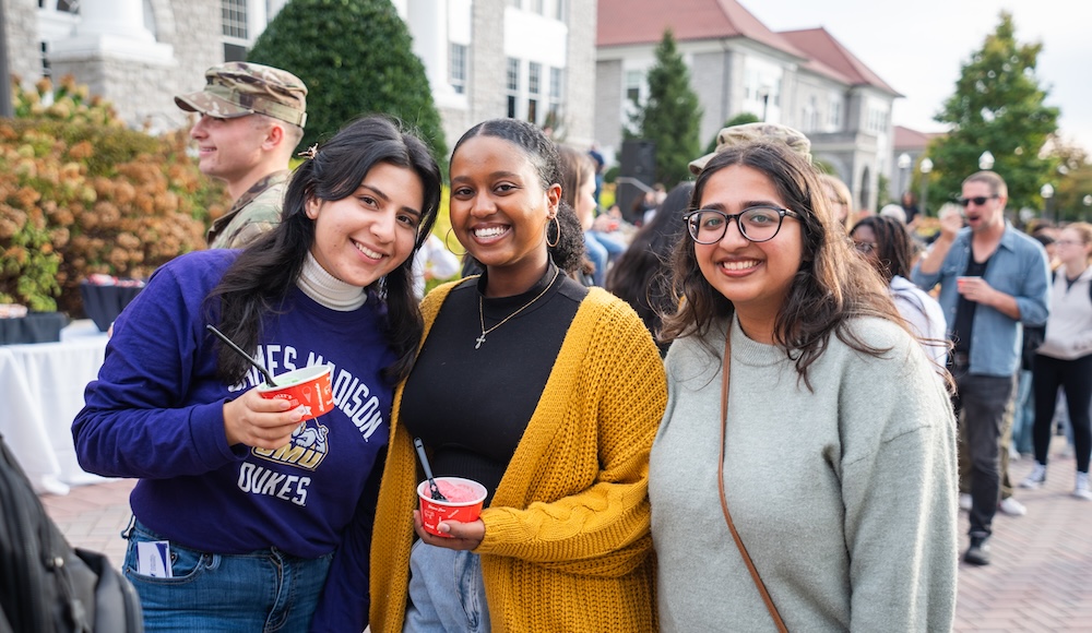 Three college students smiling and posing together while enjoying ice cream at an outdoor campus event.