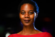 Melissa A Watkins. A Black woman with a short afro, wearing a red sweater, seen from the shoulders up against a black background.