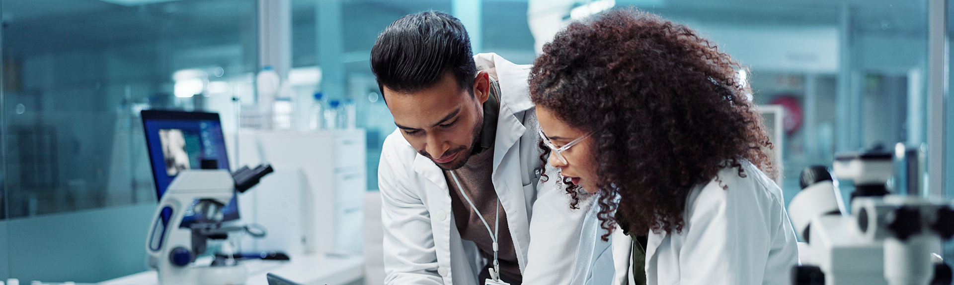 Two people are standing in a scientific laboratory, wearing lab coats, and looking at information on a tablet.