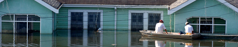 two men in a boat in flood waters; there is a single-story wood-frame house behind them; the floodwater level is about halfway up the walls of the house