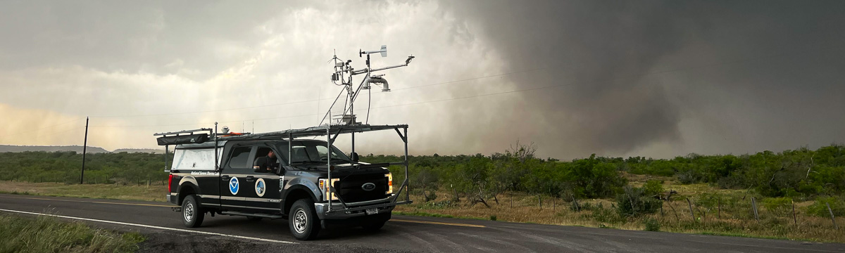 Truck with weather observation equipment mounted to the roof with tornado in the background