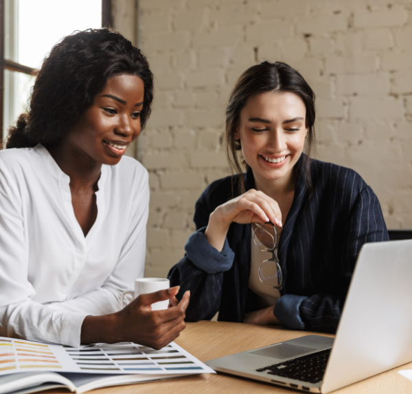 Two women sitting side by side and smiling over a laptop.