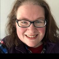 A smiling woman with long, curly brown hair and glasses, wearing a purple jacket and a multicolored shirt. She poses in front of a white background.