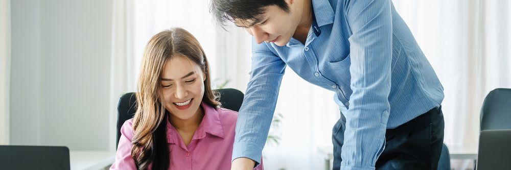 A man in a blue shirt leans over to assist a smiling woman in a pink blouse who is using a laptop in a brightly lit office.