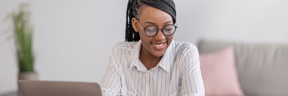 A smiling woman with braided hair and glasses working on a laptop in a bright, modern room.