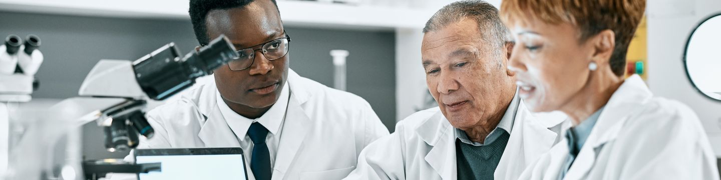 Three scientists in lab coats focus intently on their project in a laboratory. A microscope and a laptop, essential tools for their forensic psychology research, are on the table before them. One scientist works on the computer while the others observe. Shelves with scientific equipment fill the background.