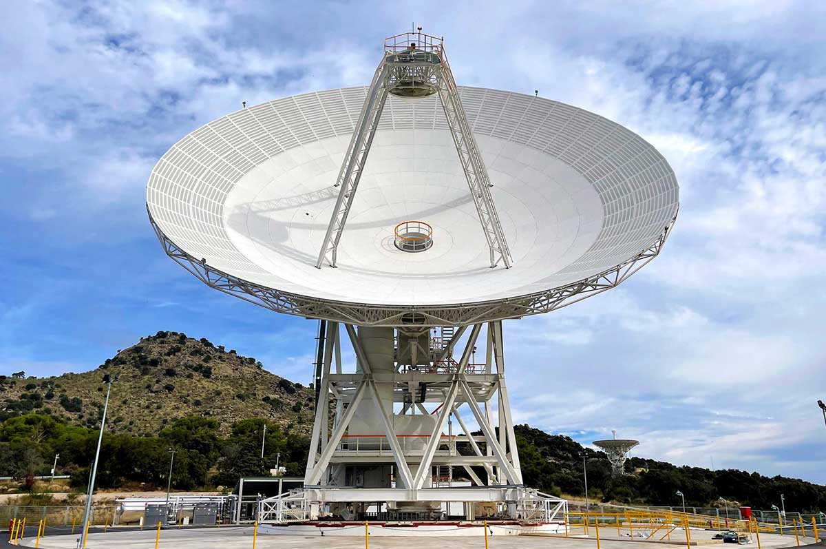 a large satellite dish is surrounded by a chain fence and other equipment with a small hill in the background. Another dish is visible among the hills.