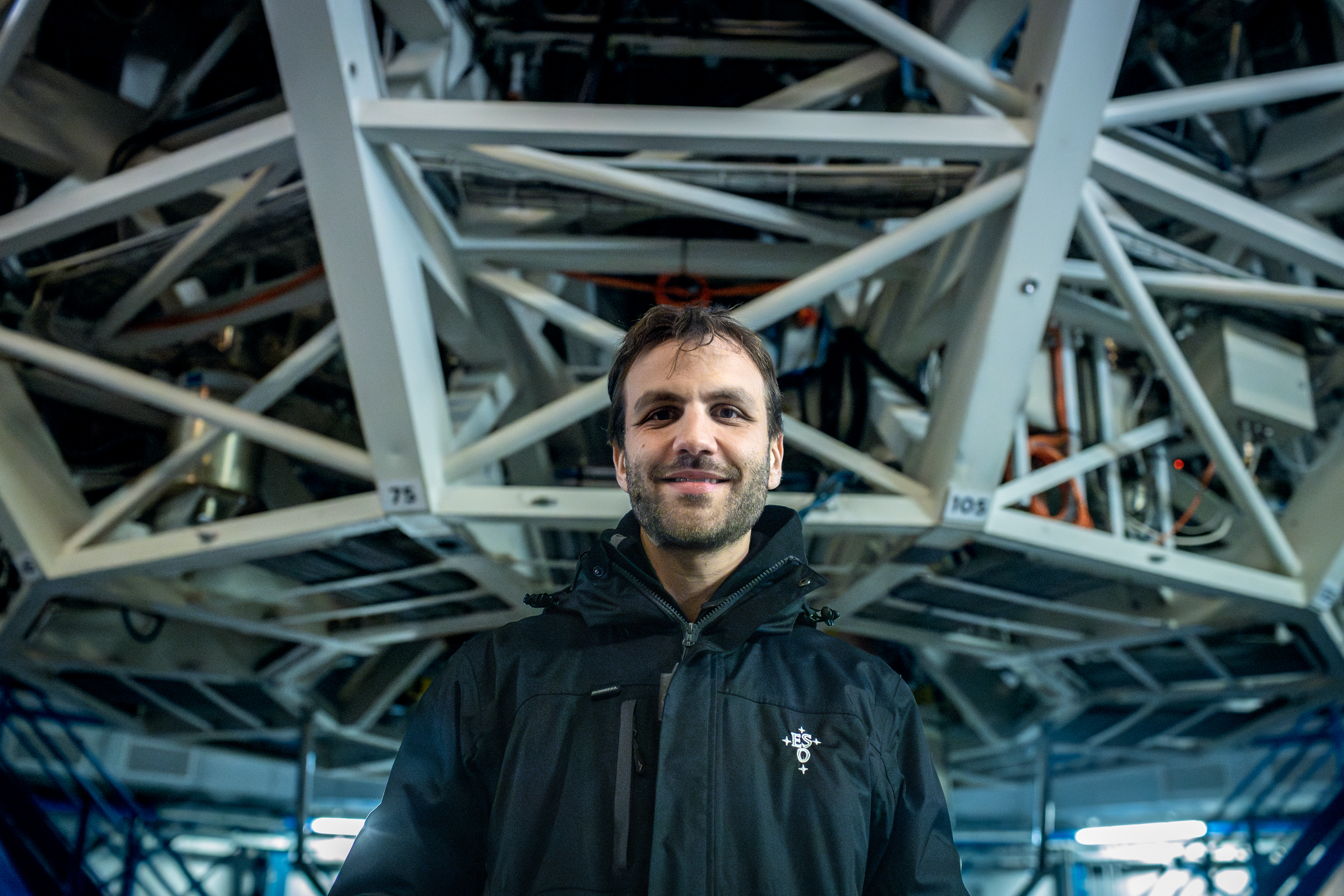 A man in a black jacket smiles at the camera, with large interlocking metal beams behind him.