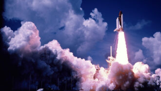 Boosted by rocket propellants, a rocket blasts off carrying the space shuttle Discovery. Clouds of smoke and steam billow near the ground.