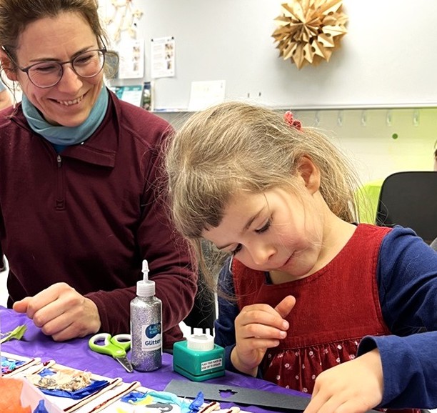 A child sitting at a table with a smiling adult, working on a craft project using paper, scissors, tape and glue.