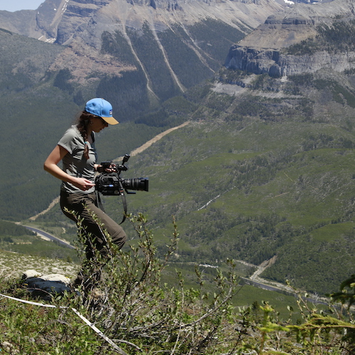 Woman with had on stands on top of a mountain with camera.