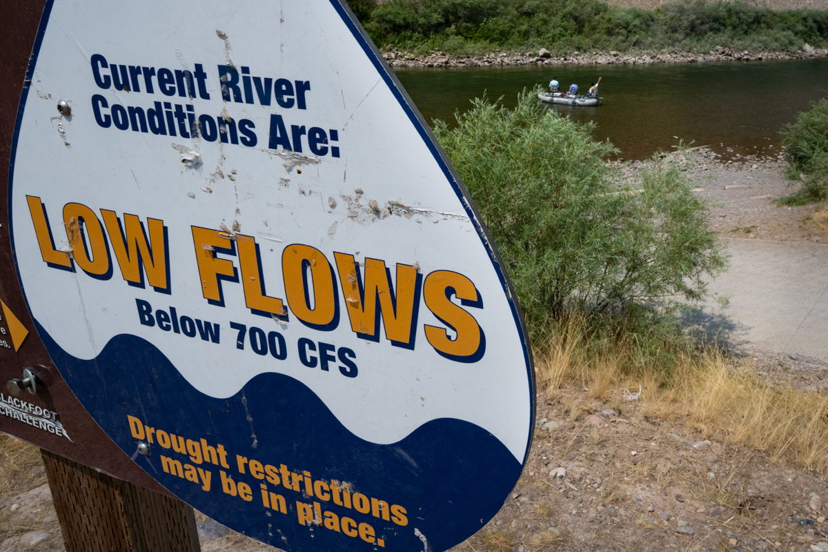 A sign near a fishing access point on the Blackfoot River warns users of low flows while a group of people fish from a raft nearby.