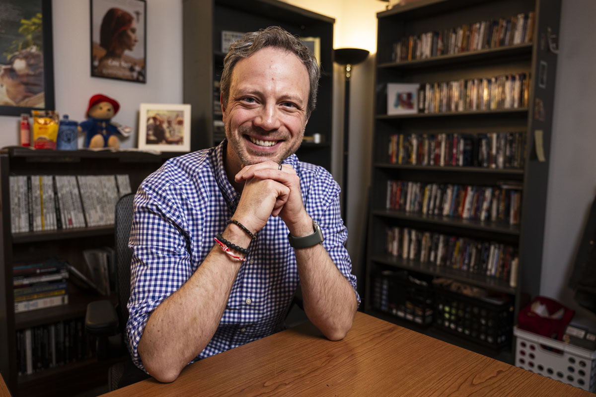 University of Montana Media Arts Adjunct Professor Tobin Addington poses for a portrait photograph in an office with a bookshelf behind him.