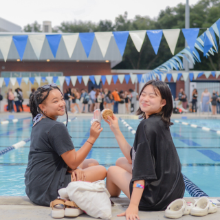 Two students sit on the side of the pool and cheers ice cream and smile