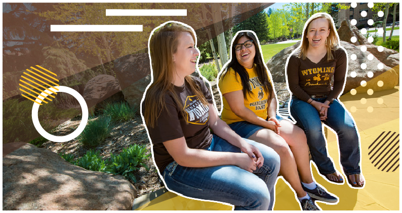 Three students in UW clothing sit together smiling against a green landscape.