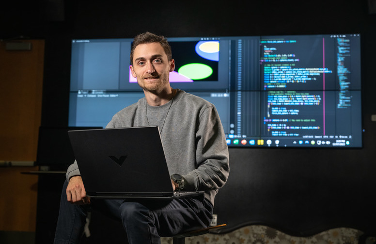 student in front of many computer screens with laptop on his knee